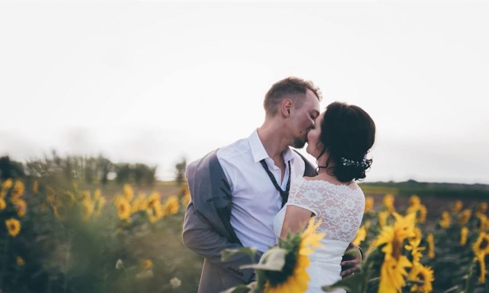 An enchanting scene featuring a couple kissing amidst vibrant sunflowers. The man is in a gray suit while the woman wears a delicate white lace dress. They embrace passionately, standing surrounded by tall sunflowers under a sky lit with soft sunset colors. Their love shines through this serene setting giving an intimate feel that showcases their connection beautifully.