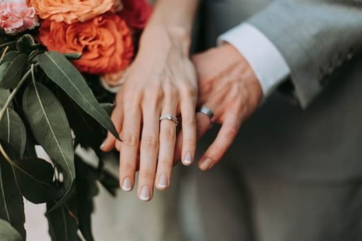 The image shows two hands wearing beautiful wedding rings together with colorful flowers in the background. One hand has part of a grey suit sleeve visible, highlighting an intimate moment of commitment between the couple. This romantic scene invokes love and celebration, perfect for weddings or engagements.