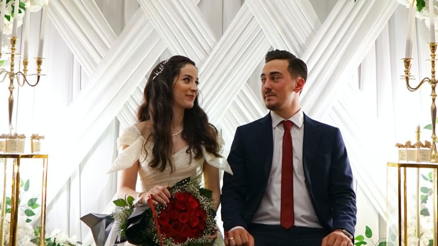 A romantic image of a bride and groom seated together at their wedding ceremony. The bride wears an elegant white dress while holding red roses and has her dark hair styled beautifully. The groom is dressed formally in a navy suit with a bright red tie, as they share happy smiles against a lovely backdrop adorned with greenery and gold candle holders, creating an enchanting atmosphere that reflects love and joy.