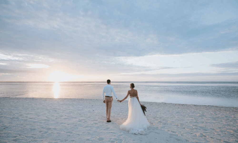 A couple walks hand in hand on a serene beach during sunset. The bride’s flowing white gown contrasts beautifully with the groom's light-colored outfit. Soft clouds float across the sky as they approach the warm ocean water, creating an ambiance of romance and tranquility perfect for weddings or intimate moments. The wide sandy beach surrounds them while the colors of sunset illuminate this peaceful scene inviting couples to create their own wedding dream here in Dubai.