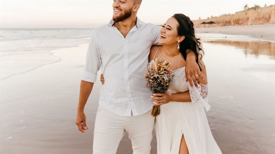 A couple stands happily on a sandy beach as waves gently lap at the shore. The man wears a white shirt with light pants while his partner is dressed in a flowing white gown adorned with lace sleeves. She holds a beautiful bouquet of flowers. They smile at each other, basking in warm sunlight against an idyllic ocean backdrop filled with calm waves and distant cliffs, creating an atmosphere of joy and tranquility perfect for their wedding day celebration.