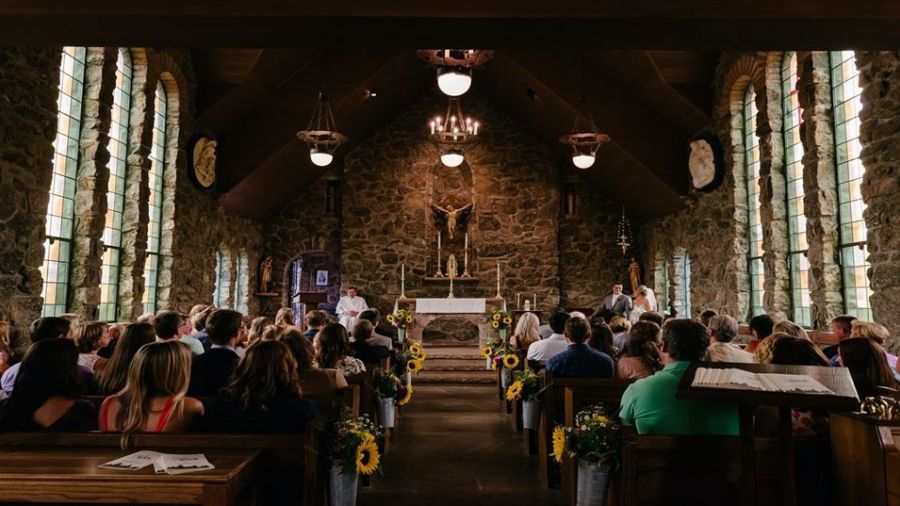 The image shows a lovely church filled with guests enjoying a wedding ceremony. Sunflowers line the aisle while light streams through arched windows enhancing the joyous atmosphere. An officiant stands at an altar adorned with candles symbolizing love and unity making this a heartwarming moment for everyone involved.
