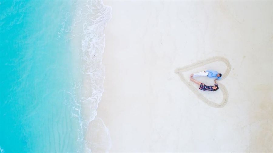 A couple lying together forms a heart shape on a sandy beach with turquoise ocean waves nearby. The man wears a light blue shirt and white pants while the woman is dressed in a dark floral dress. They appear relaxed and happy surrounded by powdery sand under bright sunlight.