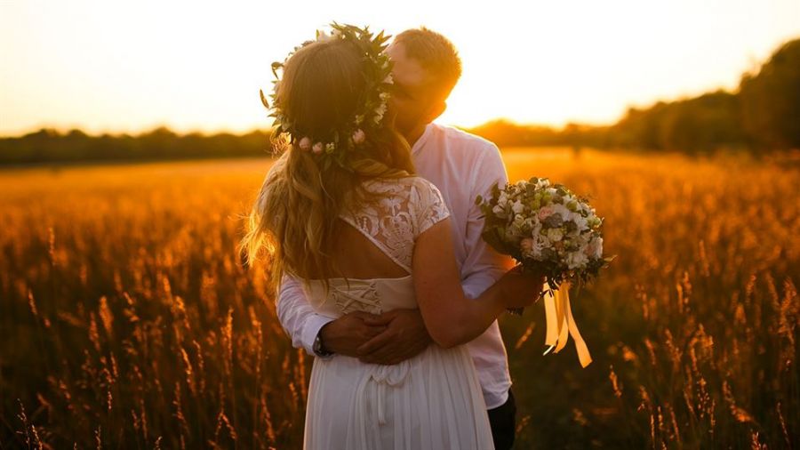 A couple embracing at sunset in a golden field, surrounded by tall grasses. The woman wears a delicate white dress and floral crown, holding a bouquet of flowers while the man dons a casual white shirt. The warm glow from the sun illuminates their joyful expression, creating an intimate atmosphere amidst soft silhouettes of trees against colorful skies.