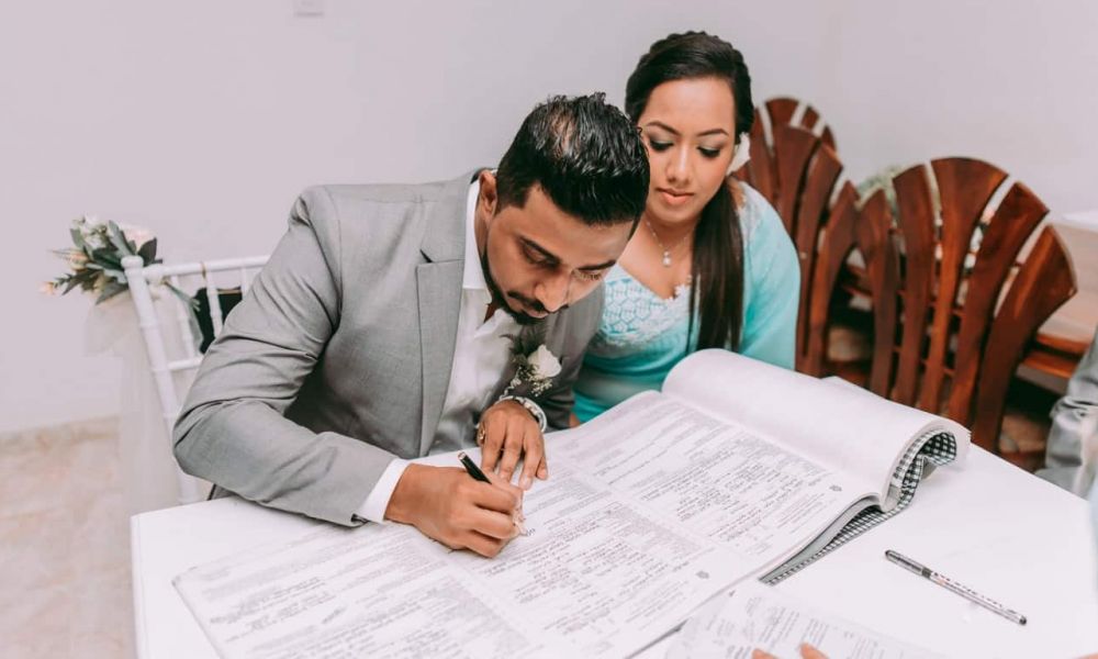 An image of a couple at their wedding ceremony shows them preparing to sign important documents. The man wears a gray suit while seated next to a woman in a light blue dress. They sit at a formal white table that holds an open ledger representing official documentation. The scene is set with minimal floral decor, creating an intimate atmosphere as they celebrate their love together through marriage.