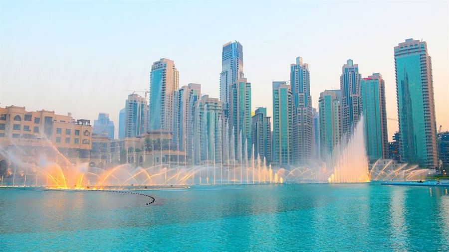 A beautiful sunset over the Dubai skyline showcases tall modern buildings reflecting on water pools. A colorful fountain show lights up the scene, creating a magical atmosphere perfect for weddings. The sparkling waters dance harmoniously as skyscrapers stand proudly in the background, displaying Dubai's architectural wonders. Ideal for couples dreaming of getting married or celebrating their love amidst stunning views, this vision captures romance and excitement that awaits you in Dubai.