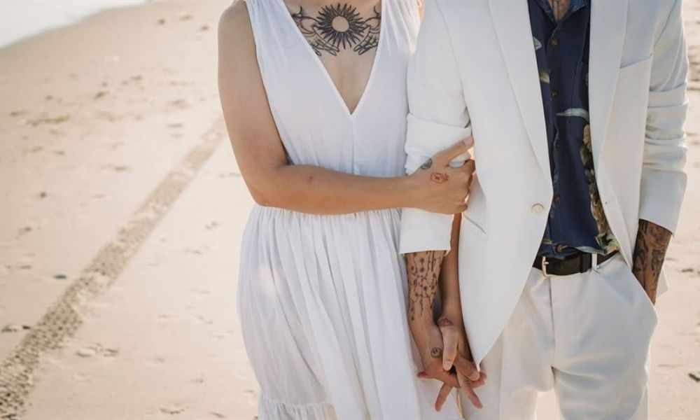 Easy Wedding in Seychelles for Dubai expatriates e58b9669 | Easy Wedding in Seychelles for Dubai expatriates | Read on, let me show you how easy wedding in Seychelles is possible as an expat in Dubai. A happy couple walks hand-in-hand on a sandy beach showcasing a romantic moment. One is dressed in an elegant sleeveless white dress while the other wears a stylish white suit with a dark shirt. Both partners display unique tattoos adding personal flair as they enjoy their special day together near the water's edge with tire tracks visible in the sand suggesting recent arrival or fun times ahead.