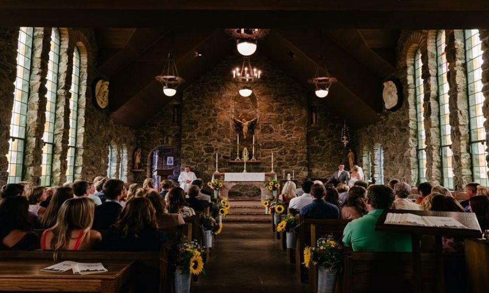 The image shows a lovely church filled with guests enjoying a wedding ceremony. Sunflowers line the aisle while light streams through arched windows enhancing the joyous atmosphere. An officiant stands at an altar adorned with candles symbolizing love and unity making this a heartwarming moment for everyone involved.