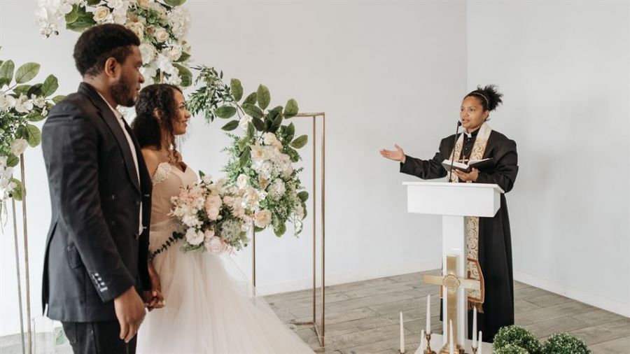 The image shows a joyful couple holding hands during their wedding ceremony in an elegant setting filled with floral arrangements. They appear happy as they stand before an officiant who conducts the ceremony in a warm atmosphere, highlighting love and commitment.
