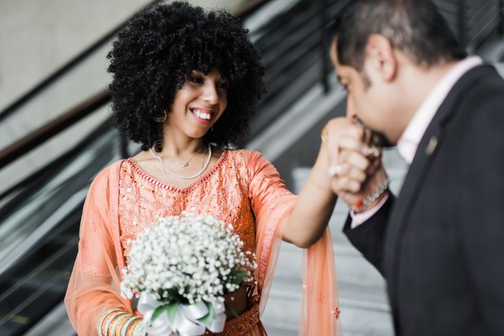 A joyful couple stands on stairs celebrating their engagement or wedding moment together. The woman wears an elegant peach dress with beautiful embroidery while holding a bouquet of white flowers. The man in dark suit kisses her hand displaying affection against a festive backdrop creating a memorable scene filled with love.