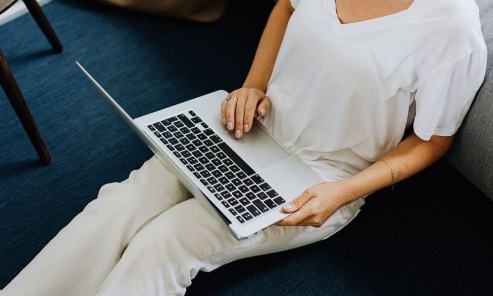 A person sits relaxed on a floor with their back against a couch while engaged with an open laptop on their lap. They wear a white T-shirt and light-colored pants, in a room with dark blue carpet that contrasts nicely with their outfit. The subtle lighting creates a calm atmosphere, making it look like they are working or enjoying leisure time at home under natural light.