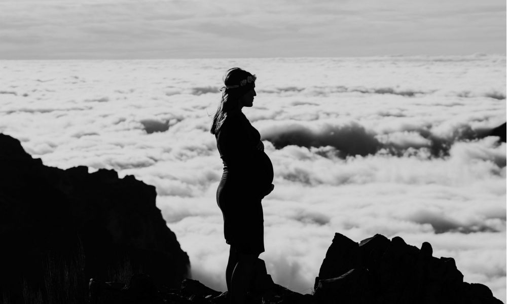 A silhouette of a pregnant woman in a dress stands on a rocky cliff, looking out over clouds. The scene captures her profile against a vast sky filled with fluffy white clouds. The contrast of darkness from the rocks and brightness from the clouds creates an emotional tone, portraying tranquility and reflection as she enjoys nature's beauty around her while contemplating life ahead.