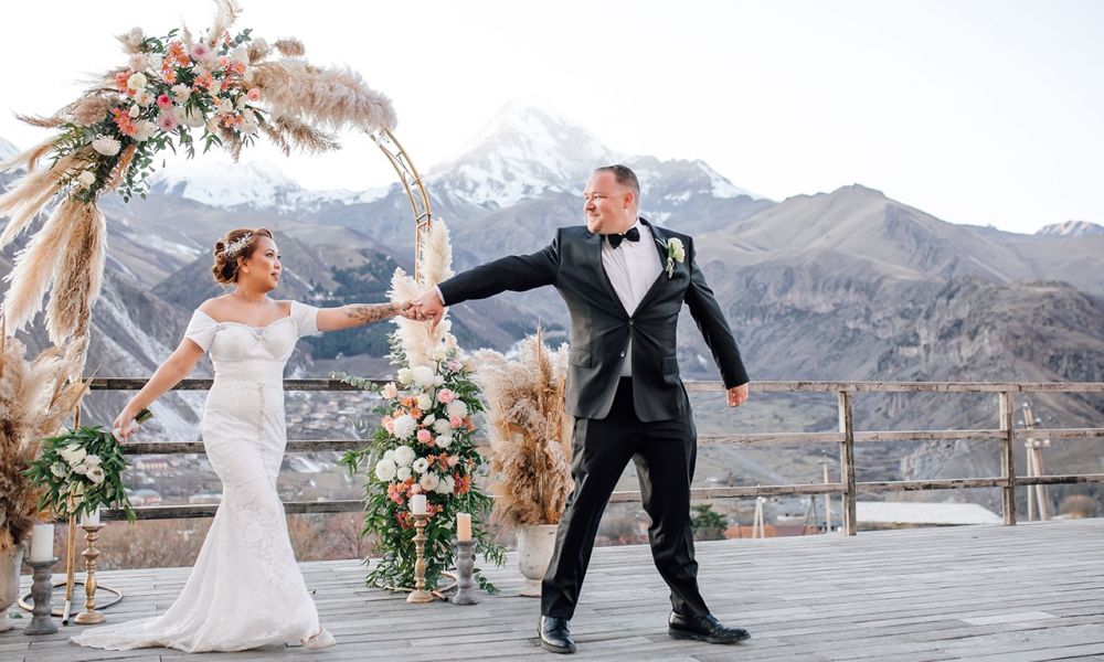 will uae expats find winter wedding in georgia cheaper 1 ec92b067 | Winter Wedding in Georgia vs. Dubai: A Budget-Friendly Dream for UK and UAE Expats | Planning a winter wedding in Georgia can be the perfect alternative for expats living in the UAE and couples from the United Kingdom. While Dubai is often seen as a glamorous place to get married, the high prices can be overwhelming. Georgia offers a charming and affordable option—especially during the winter months. A happy couple stands outdoors on a wooden platform for their wedding ceremony. The bride wears an elegant white lace gown with a floral headpiece while the groom is dressed in a classic black tuxedo. Behind them, there is an arch of flowers and pampas grass framing the majestic snow-covered mountains under a bright blue sky creating a romantic atmosphere for their special day.