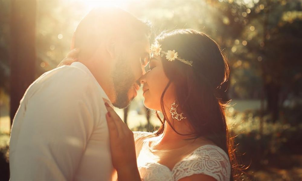 The image shows a couple warmly embracing outdoors in soft sunlight. The man wears a white shirt while the woman is dressed in a beautiful lace gown adorned with flowers. Their joyful expressions portray love and closeness, surrounded by blurred greenery that adds serenity to the scene. The lighting creates a dreamy effect hinting at a magical moment just before their kiss. This romantic setting captures the essence of weddings in Dubai and Abu Dhabi beautifully.