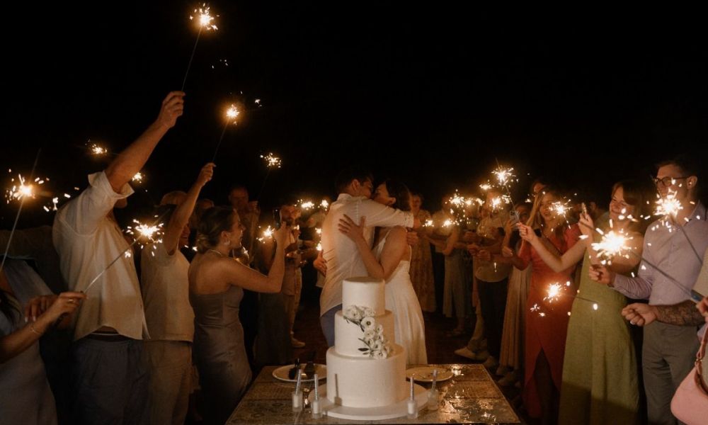 A joyful couple embraces at their wedding celebration in Dubai. They stand before a beautiful white wedding cake decorated with flowers while guests surround them holding sparklers that light up the night sky. The guests are dressed elegantly, smiling as they celebrate this special moment together, creating a warm atmosphere filled with love and happiness.