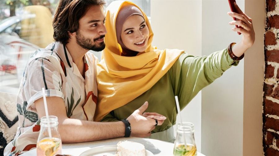 A happy couple sits at a brightly lit table enjoying drinks together. The woman is wearing a colorful headscarf and green top while holding a phone as if taking a selfie. The man has medium-length hair and a patterned shirt. They look relaxed smiling at each other in a cozy dining spot with brick walls, creating an inviting atmosphere perfect for sharing joyous moments.