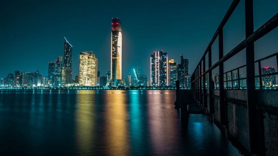 The image features a breathtaking nighttime view of Abu Dhabi with illuminated skyscrapers reflecting on water. It shows a vibrant cityscape filled with lights creating an enchanting atmosphere. A walkway extends over the tranquil surface, leading into this lively scene where modernity meets natural beauty under deep blue skies accented by warm yellow lights.