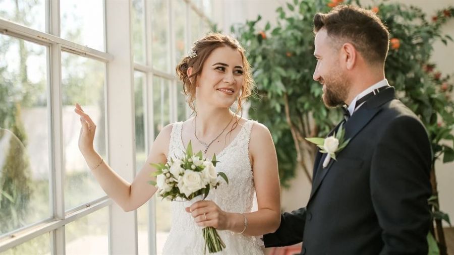 An excited couple stands inside a bright room adorned with large windows and lush green plants. The bride wears a gorgeous white lace wedding dress, holding a bouquet of white flowers while smiling radiantly at her partner. He looks dapper in a dark suit with a bow tie. The light streaming through the windows creates an enchanting atmosphere for their special day.