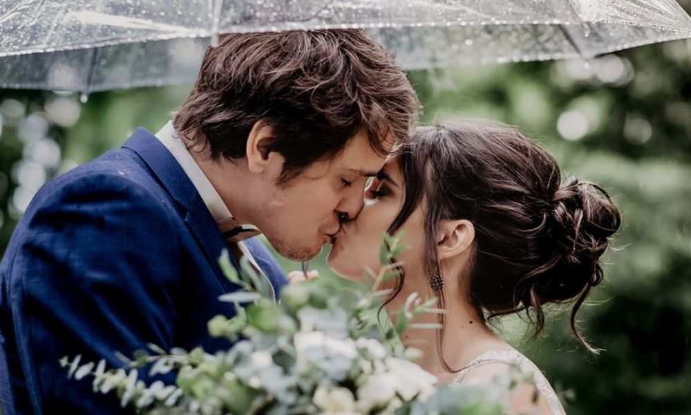 A couple sharing a kiss under a transparent umbrella in a lush green setting represents love and togetherness. The man wears a dark blue suit while the woman is in an elegant white gown. They are surrounded by nature as light rain falls around them creating a romantic atmosphere. The woman's bridal bouquet features various green and white flowers which adds beauty to this intimate moment captured outdoors.