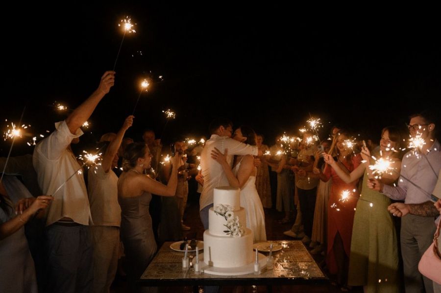 A joyful couple embraces at their wedding celebration in Dubai. They stand before a beautiful white wedding cake decorated with flowers while guests surround them holding sparklers that light up the night sky. The guests are dressed elegantly, smiling as they celebrate this special moment together, creating a warm atmosphere filled with love and happiness.