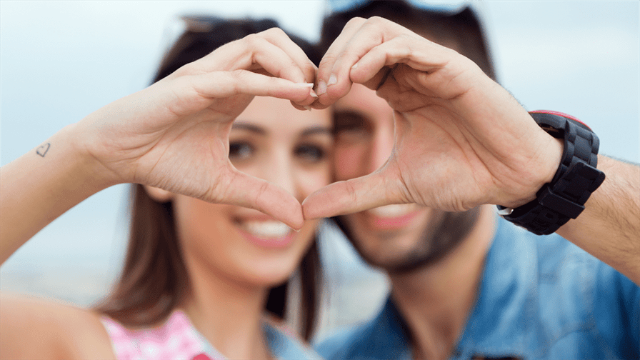 The image shows a happy couple forming a heart shape with their hands against a blurry background. They are smiling and casually dressed, radiating joy and love. The woman has long hair with sunglasses on her head while the man also has sunglasses pushed back. This romantic moment emphasizes the beauty of connection between them as they celebrate their love story in Dubai.