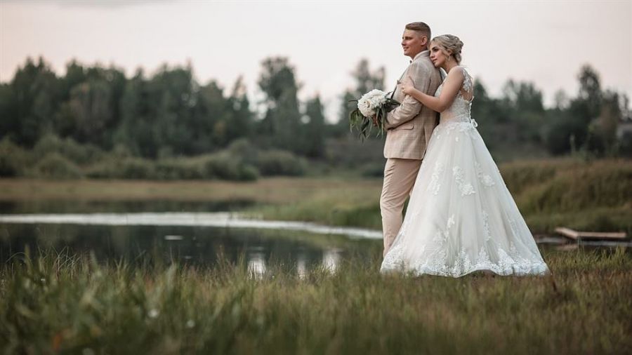 A lovely couple stands by a serene lake, surrounded by lush greenery. The bride wears an elegant white gown adorned with lace, while the groom looks dapper in a beige suit. They stand peacefully, with the bride gently embracing the groom from behind, holding a bouquet of white flowers. The background features soft-focus trees and a cloudy sky which adds to the romantic feeling of this tranquil scene. This captures their tender moment on their special day amidst nature's beauty.